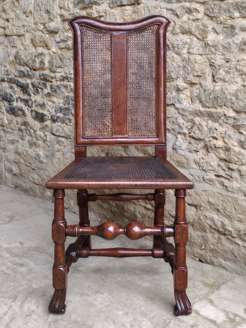 An early 18th-century dark walnut side chair featuring a high back with woven cane panels and central wooden splat, a caned seat, and turned legs with carved feet.