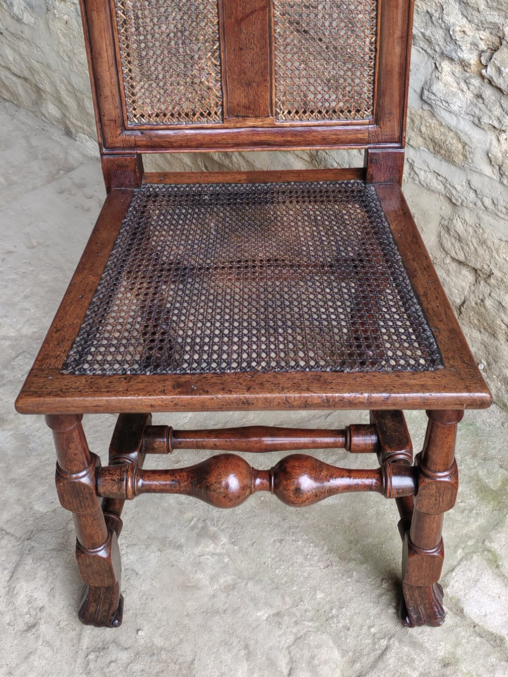 An early 18th-century dark walnut side chair featuring a high back with woven cane panels and central wooden splat, a caned seat, and turned legs with carved feet. View VI