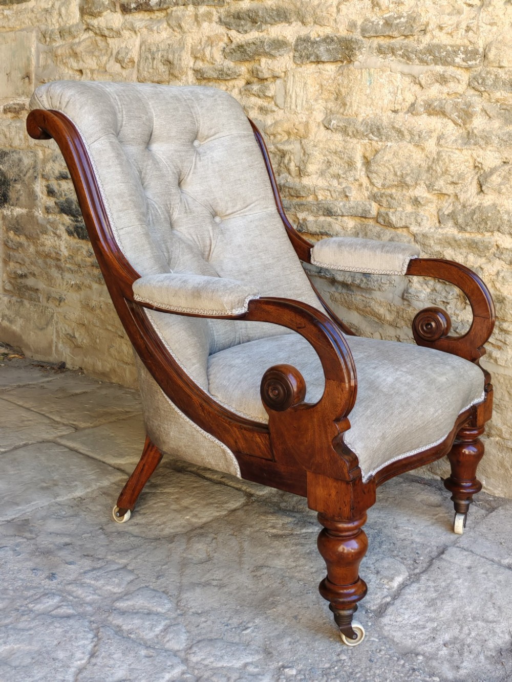 Mid-Victorian walnut armchair featuring light grey button-back upholstery, carved scroll armrests, and turned legs with ceramic casters.