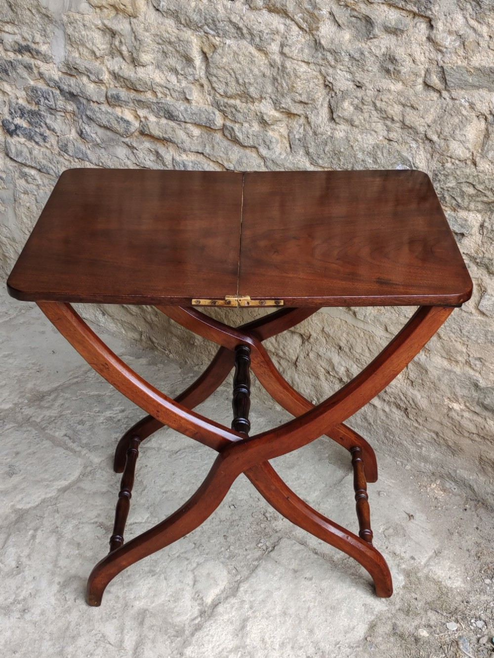 An antique mahogany coaching table featuring a hinged folding top, cross-framed legs, and turned stretchers set against a stone wall.