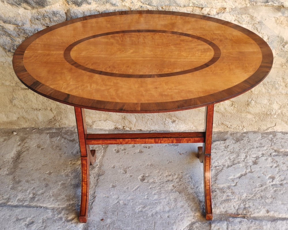 An oval satinwood coffee table featuring double band inlay on the tabletop and supported by twin trestle legs connected by a central stretcher.