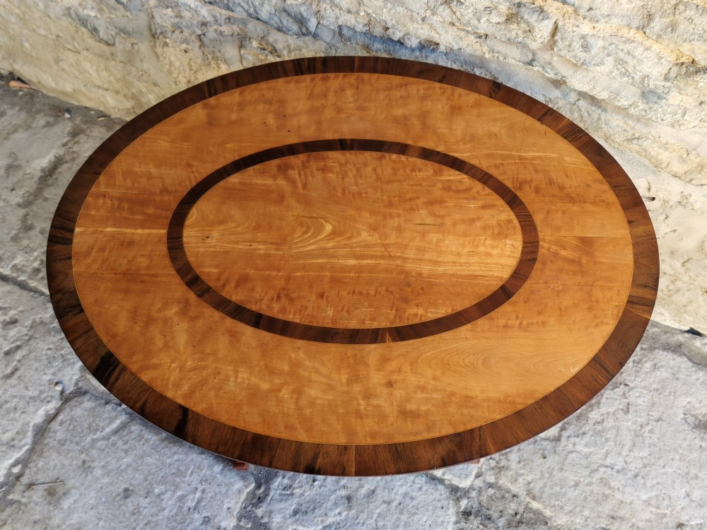 An oval satinwood coffee table featuring double band inlay on the tabletop and supported by twin trestle legs connected by a central stretcher. View II