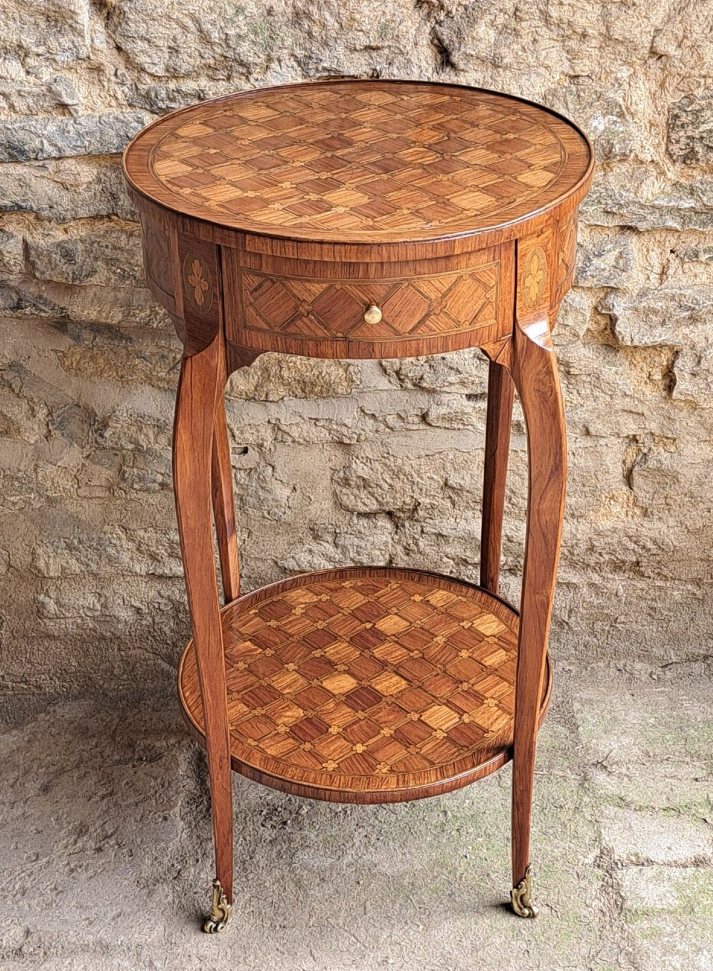 An oval two-tier kingwood occasional table featuring geometric parquetry inlay, a single drawer with a brass knob, curved cabriole legs, and brass foot sabots.