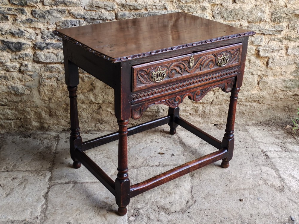 An 18th-century dark oak lowboy featuring a single relief-carved drawer with brass handles, turned front legs, and a shaped carved apron. View VI