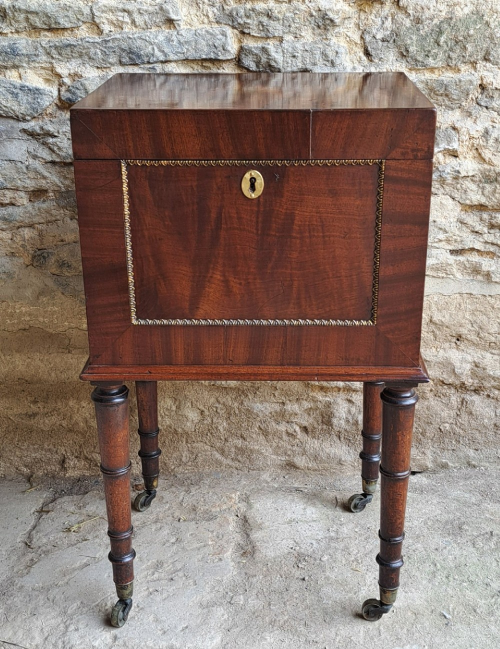 A square dark mahogany cellaret featuring a brass keyhole escutcheon, beaded ormolu border, turned legs, and brass castors.