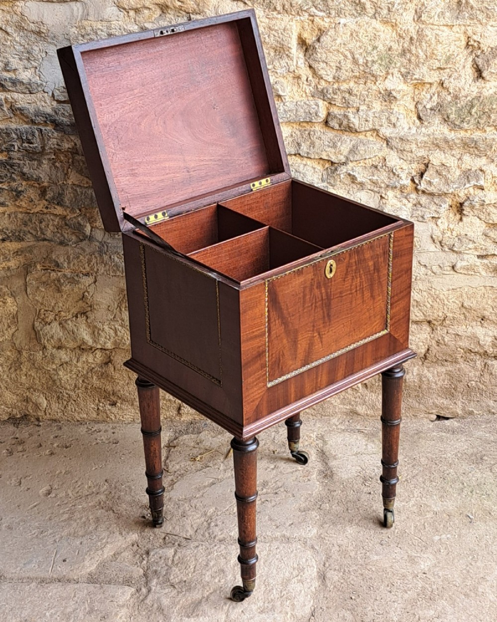 A square dark mahogany cellaret featuring a brass keyhole escutcheon, beaded ormolu border, turned legs, and brass castors. View V