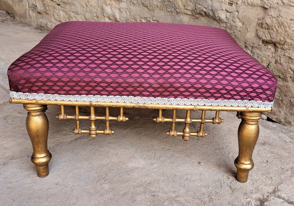 A low rectangular footstool featuring a patterned burgundy padded top with silver braid trim and a gilded wood frame with turned legs and lattice fretwork details. View V