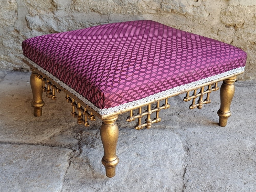 A low rectangular footstool featuring a patterned burgundy padded top with silver braid trim and a gilded wood frame with turned legs and lattice fretwork details. View VI
