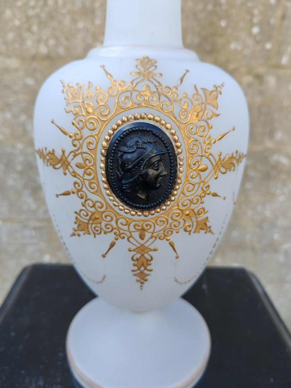 A white opaline glass vase featuring a slender neck, a pedestal base, gold filigree detailing, and a black classical profile cameo medallion at the centre. View II