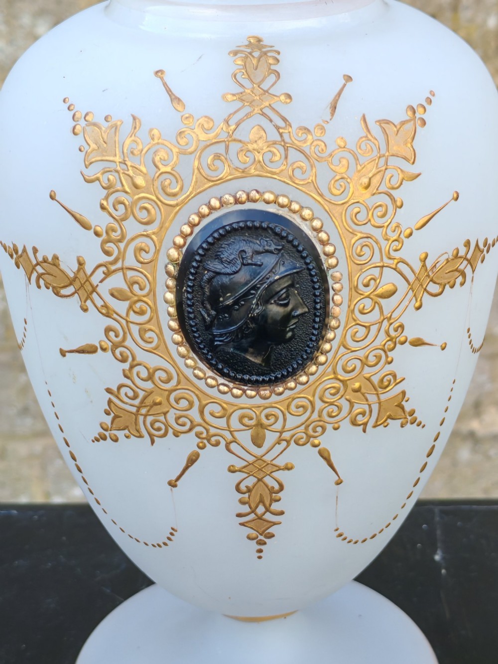 A white opaline glass vase featuring a slender neck, a pedestal base, gold filigree detailing, and a black classical profile cameo medallion at the centre. View III