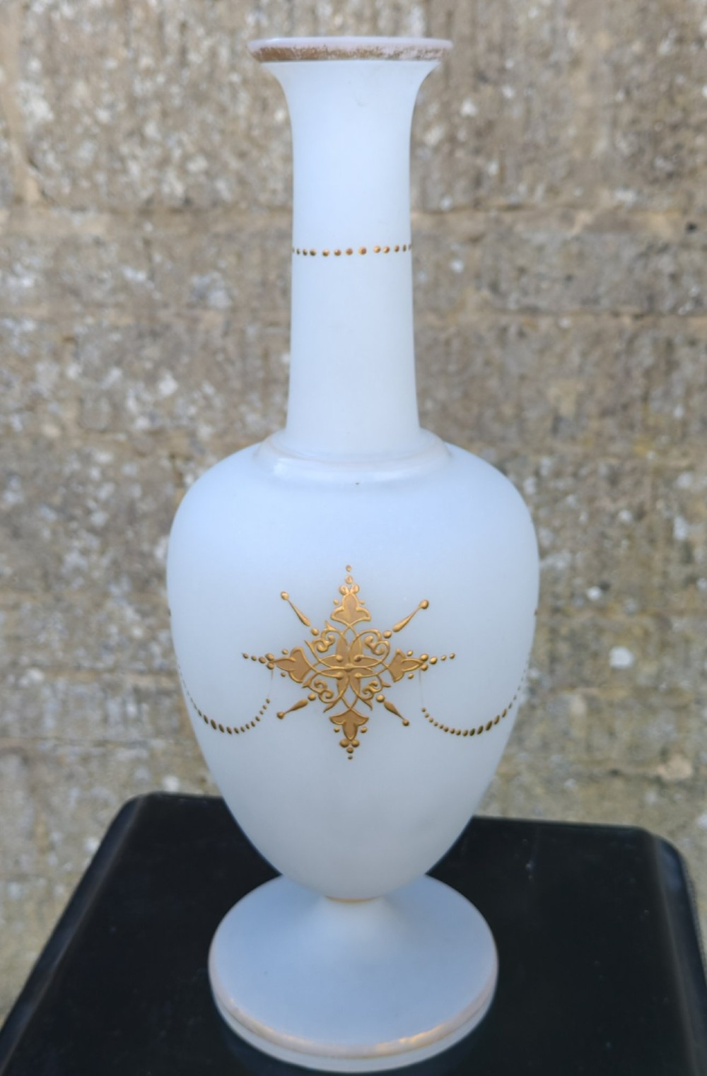 A white opaline glass vase featuring a slender neck, a pedestal base, gold filigree detailing, and a black classical profile cameo medallion at the centre. View V