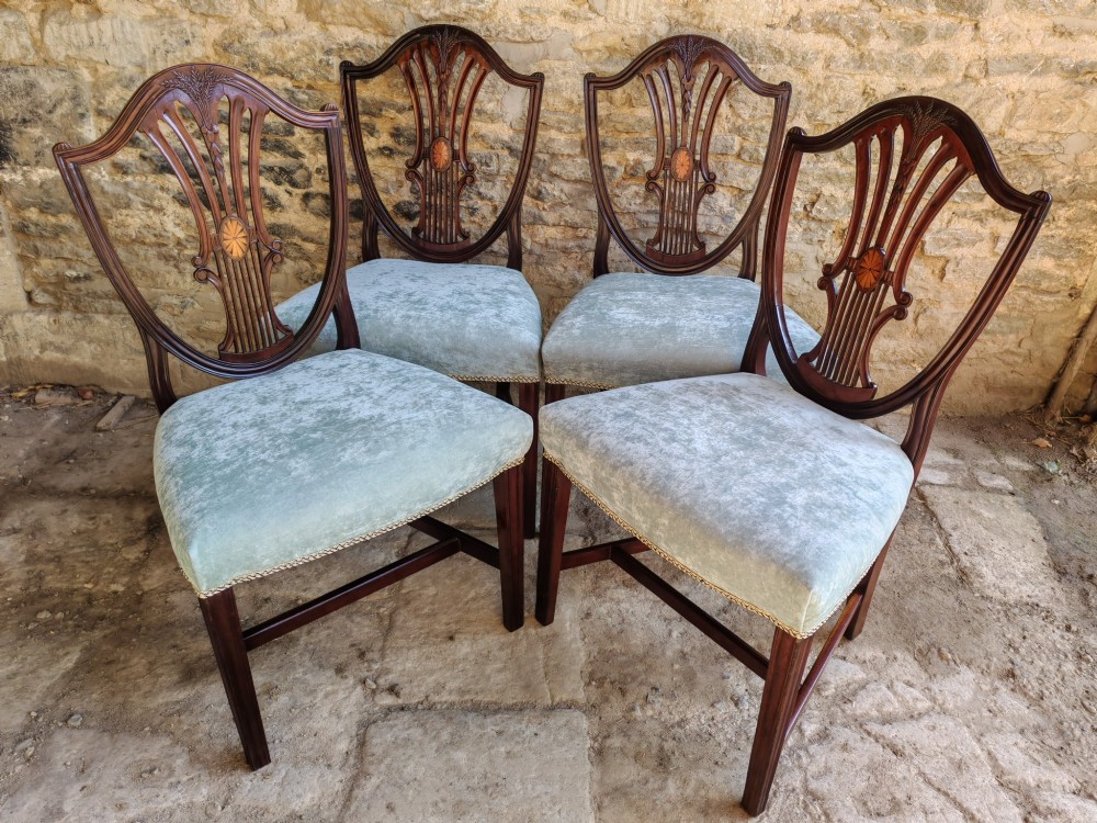 A set of four antique mahogany Hepplewhite dining chairs featuring carved shield-shaped backs with central inlay motifs and light green upholstered seats.