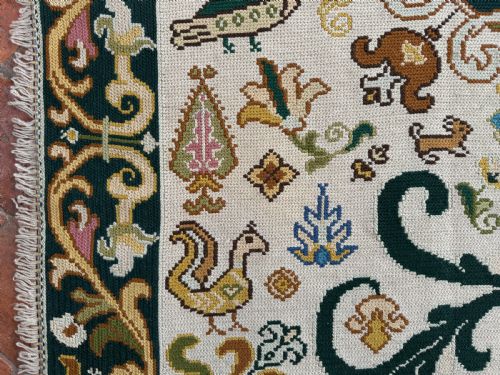 fig 2. A rectangular handstitched Portuguese needlepoint rug featuring a cream central field with scrolling foliage, bird motifs, and a dark ornate border.