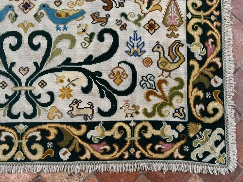 fig 4. A rectangular handstitched Portuguese needlepoint rug featuring a cream central field with scrolling foliage, bird motifs, and a dark ornate border.