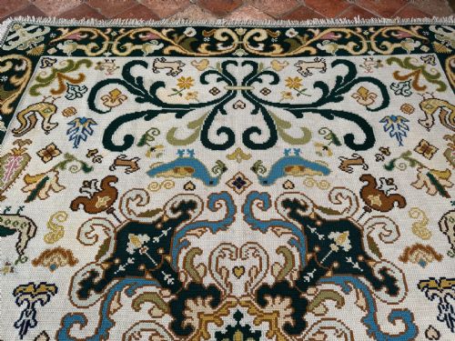 fig 5. A rectangular handstitched Portuguese needlepoint rug featuring a cream central field with scrolling foliage, bird motifs, and a dark ornate border.