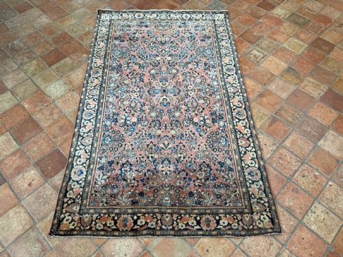 fig 1. A rectangular hand-knotted antique Sarouk rug featuring intricate floral sprays in blue and cream across a terracotta red field with a dark blue multi-banded border.