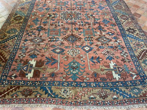 fig 1. An antique Persian Heriz hand-knotted carpet featuring a terracotta red field filled with geometric motifs, enclosed by a blue and beige patterned border.