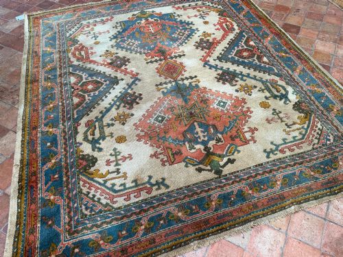 fig 1. A rectangular hand-knotted wool Ushak carpet featuring central geometric diamond medallions in terracotta red and blue set against a cream field with framed borders.