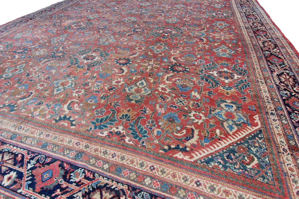 A large rectangular antique hand-knotted wool carpet featuring a red central field filled with blue and beige floral motifs bordered by dark navy outer bands. View III