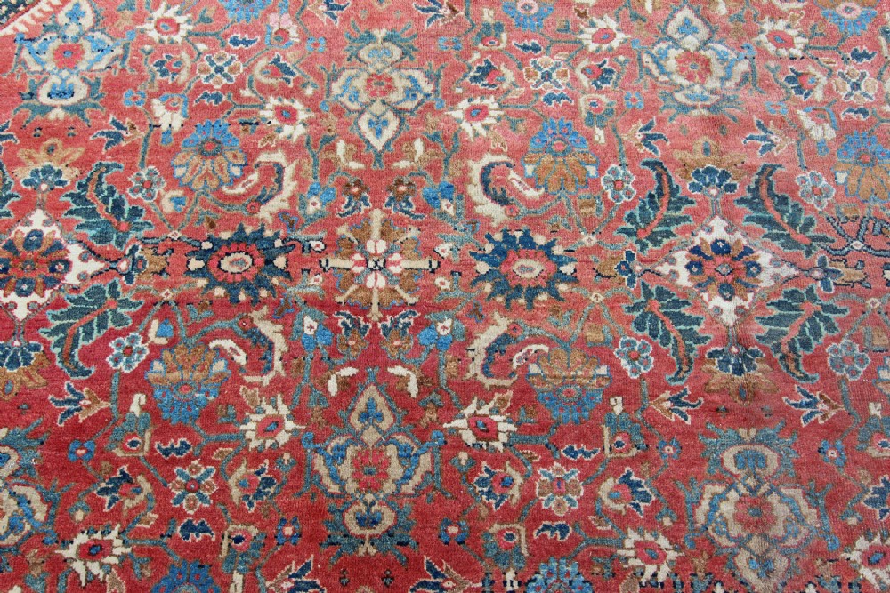 A large rectangular antique hand-knotted wool carpet featuring a red central field filled with blue and beige floral motifs bordered by dark navy outer bands. View IV