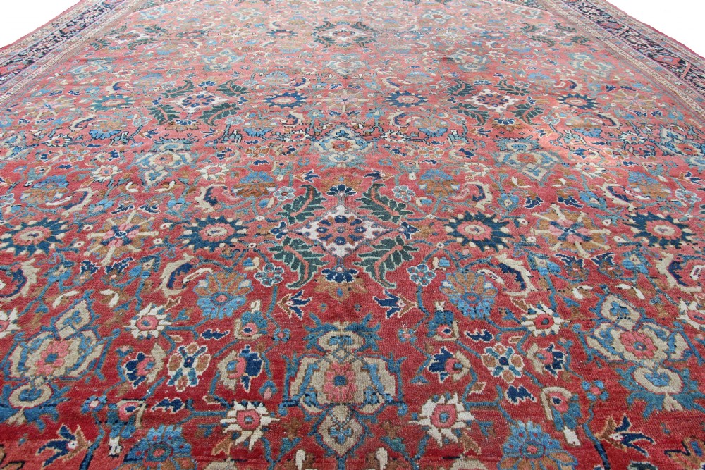 A large rectangular antique hand-knotted wool carpet featuring a red central field filled with blue and beige floral motifs bordered by dark navy outer bands. View V