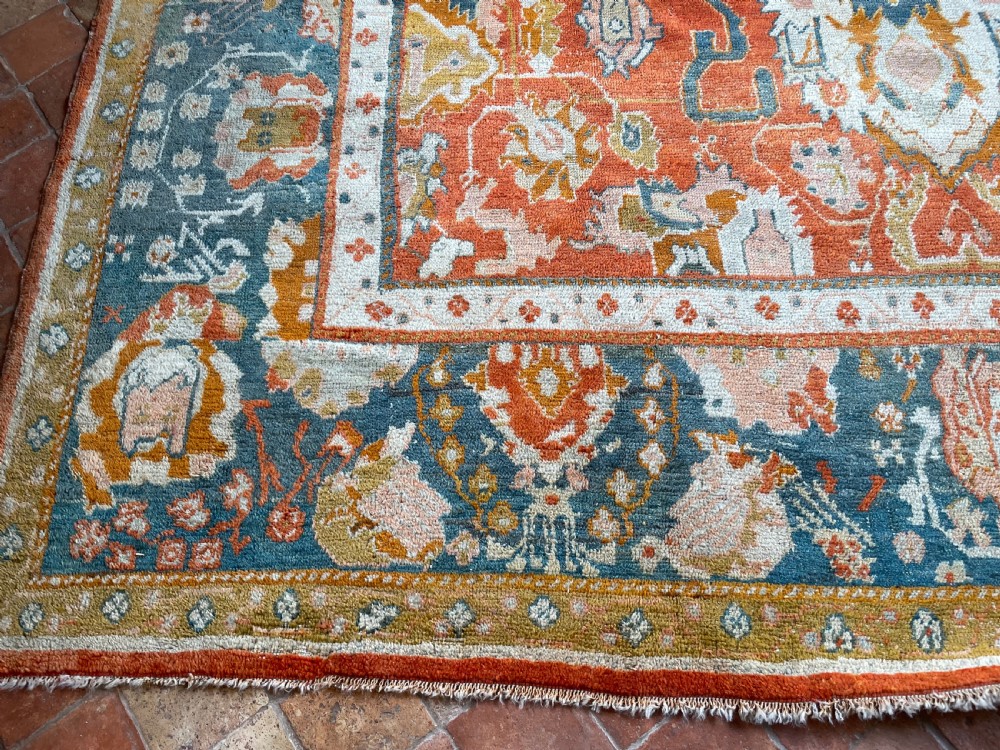 An antique hand-knotted Ushak wool carpet featuring a terracotta central field with stylised ivory medallions and a dark blue border. View V