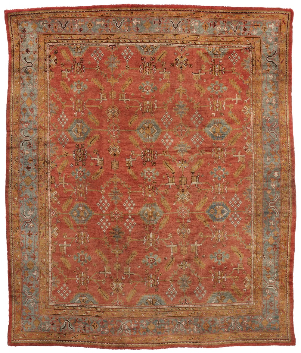 A large hand-knotted antique Ushak wool carpet featuring a terracotta field patterned with muted blue, mustard, and cream stylised geometric motifs within a layered border.