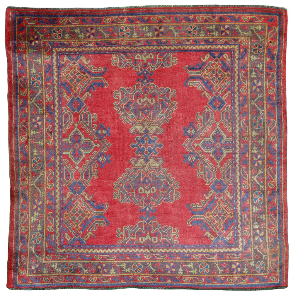 An antique square hand-knotted Ushak wool carpet featuring a vibrant red field with blue geometric central medallion motifs within a decorated multi-band border.