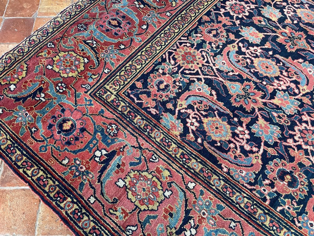 A large rectangular hand-knotted antique Sauj Bulagh carpet featuring a dark navy blue central field covered in intricate floral motifs, framed by a broad terracotta red border. View II