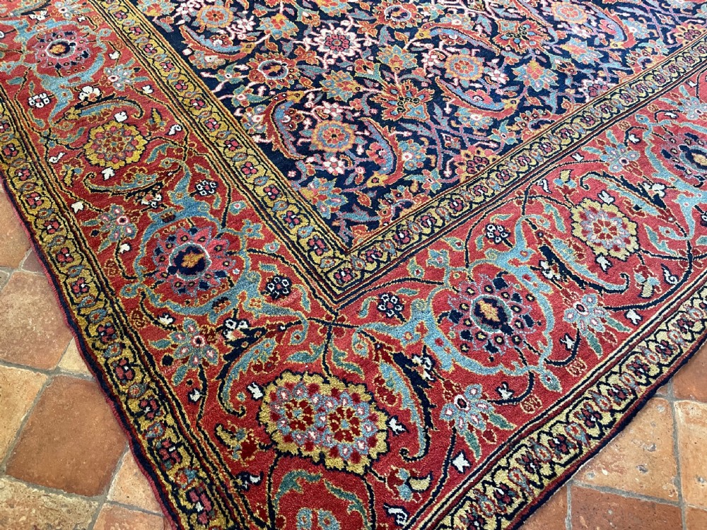 A large rectangular hand-knotted antique Sauj Bulagh carpet featuring a dark navy blue central field covered in intricate floral motifs, framed by a broad terracotta red border. View III