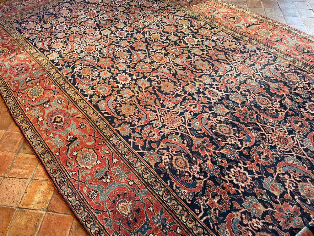 A large rectangular hand-knotted antique Sauj Bulagh carpet featuring a dark navy blue central field covered in intricate floral motifs, framed by a broad terracotta red border. View IV