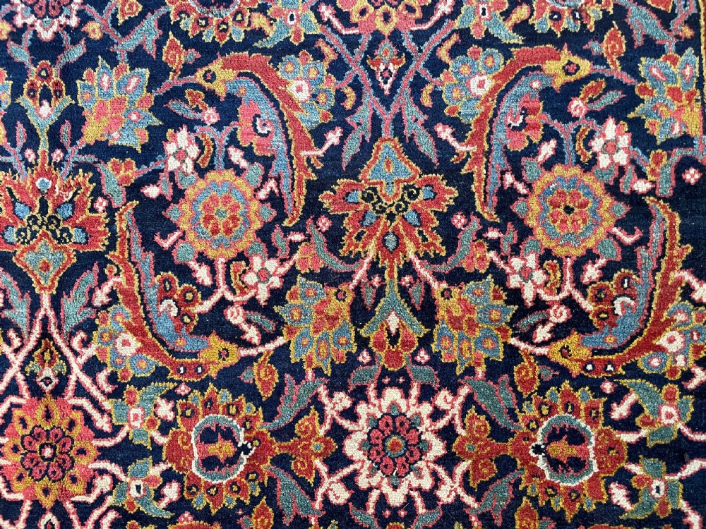 A large rectangular hand-knotted antique Sauj Bulagh carpet featuring a dark navy blue central field covered in intricate floral motifs, framed by a broad terracotta red border. View V
