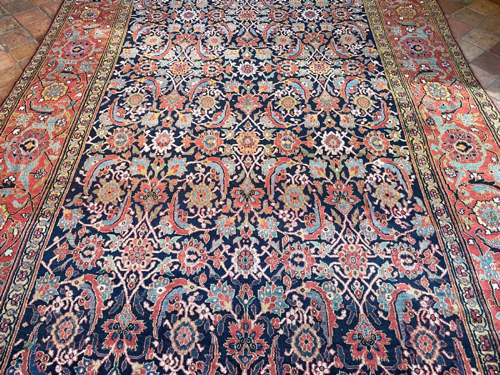 A large rectangular hand-knotted antique Sauj Bulagh carpet featuring a dark navy blue central field covered in intricate floral motifs, framed by a broad terracotta red border. View VI