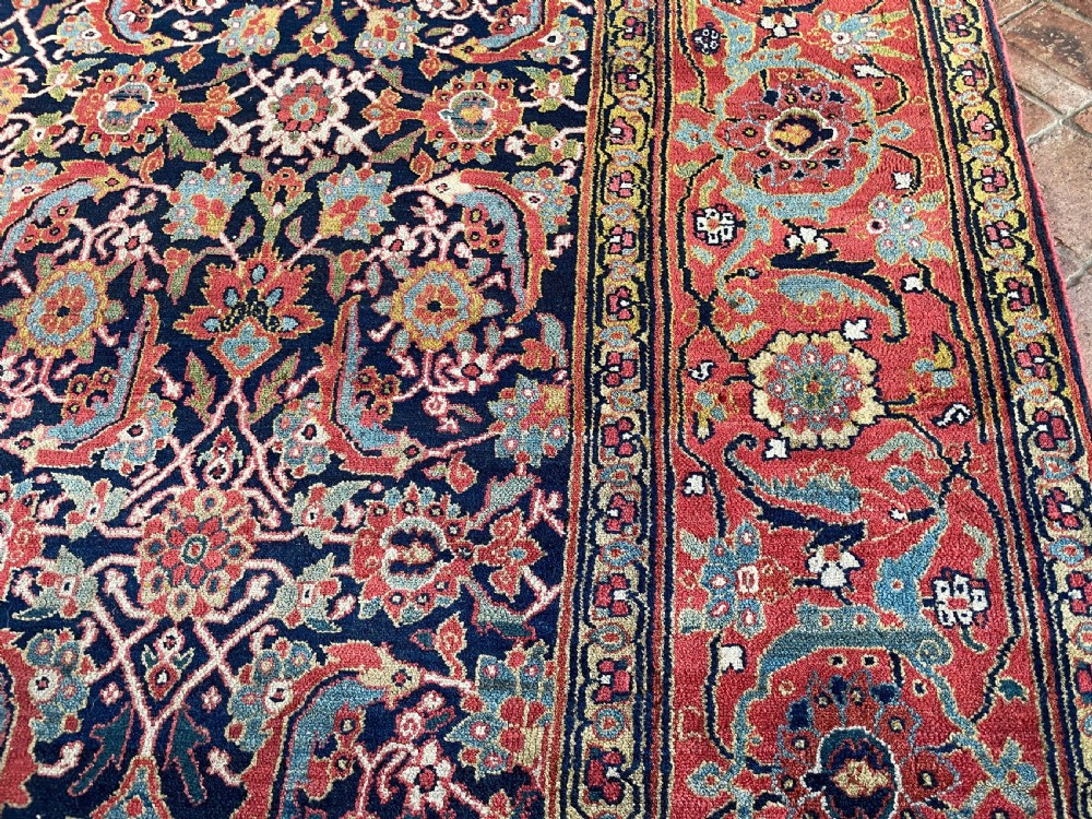 A large rectangular hand-knotted antique Sauj Bulagh carpet featuring a dark navy blue central field covered in intricate floral motifs, framed by a broad terracotta red border. View VII