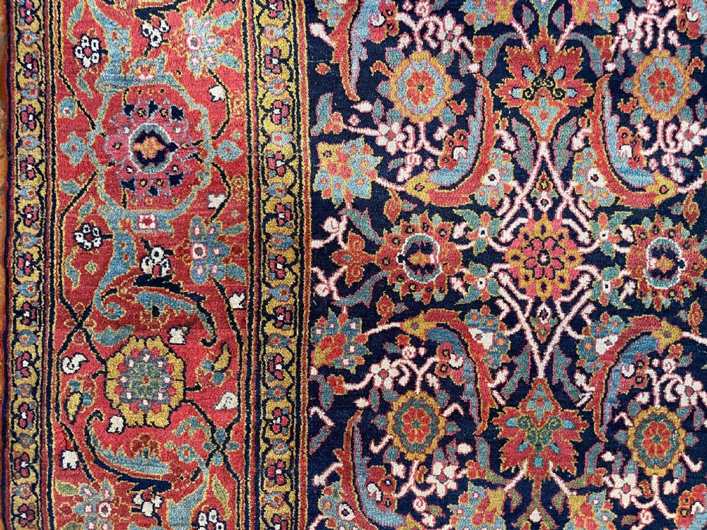 A large rectangular hand-knotted antique Sauj Bulagh carpet featuring a dark navy blue central field covered in intricate floral motifs, framed by a broad terracotta red border. View VIII