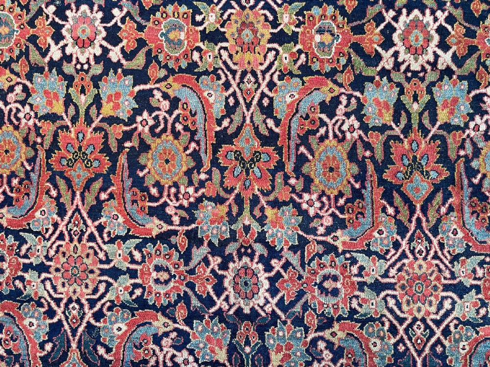 A large rectangular hand-knotted antique Sauj Bulagh carpet featuring a dark navy blue central field covered in intricate floral motifs, framed by a broad terracotta red border. View X