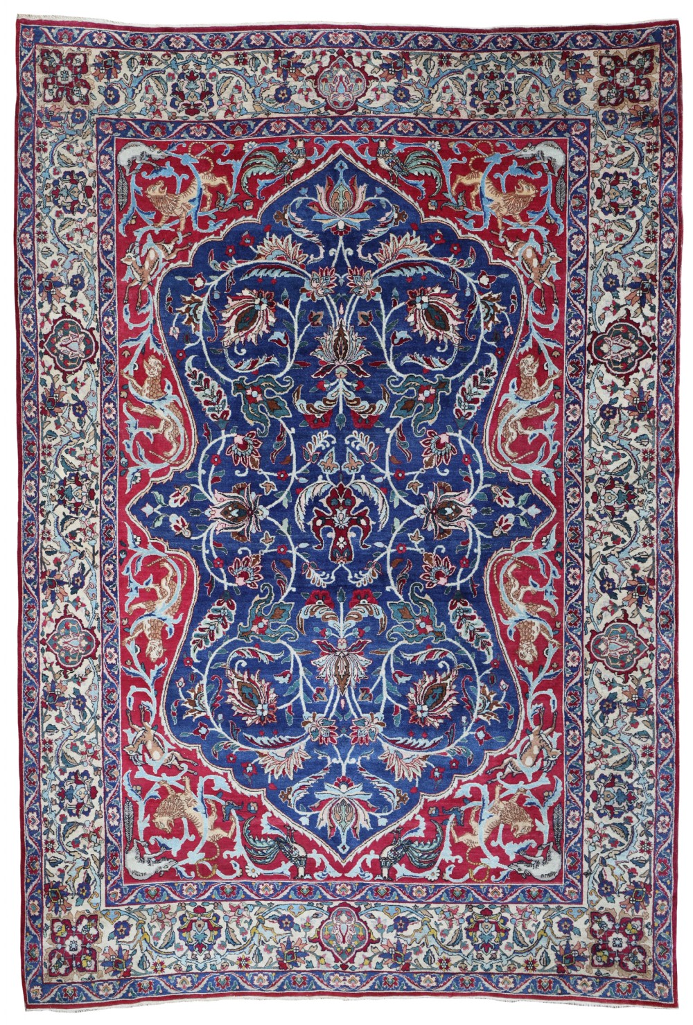An antique Persian Tehran carpet featuring a deep navy blue central field, red spandrels, and an ivory border filled with detailed floral vines and animal motifs.