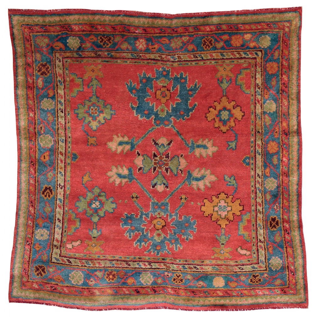 A square antique Turkish Ushak hand-knotted wool rug featuring a crimson field with teal floral medallions and a broad patterned blue border.