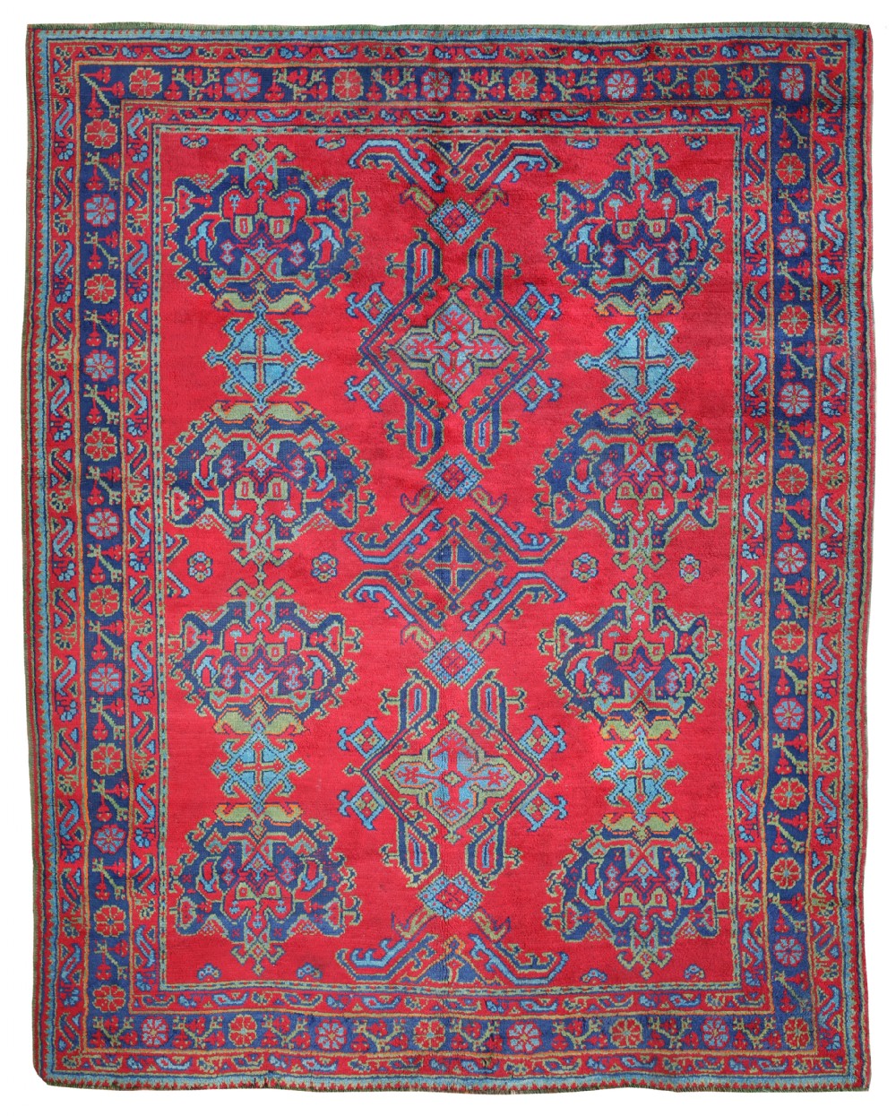 An antique Turkish Ushak wool carpet featuring a crimson red field woven with central blue geometric medallions and a matching patterned border.
