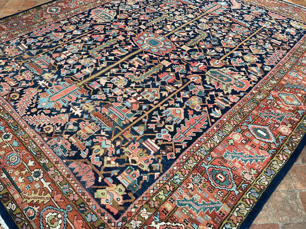 A rectangular hand-knotted antique Heriz carpet featuring a dark navy central field filled with geometric floral motifs and a wide terracotta red border. View V