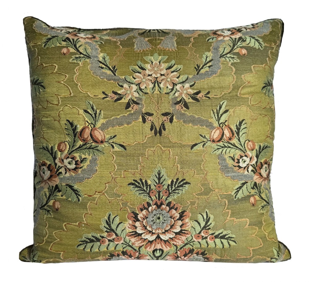 A square olive green silk cushion decorated with a woven botanical pattern in pastel pink, grey, and green tones.