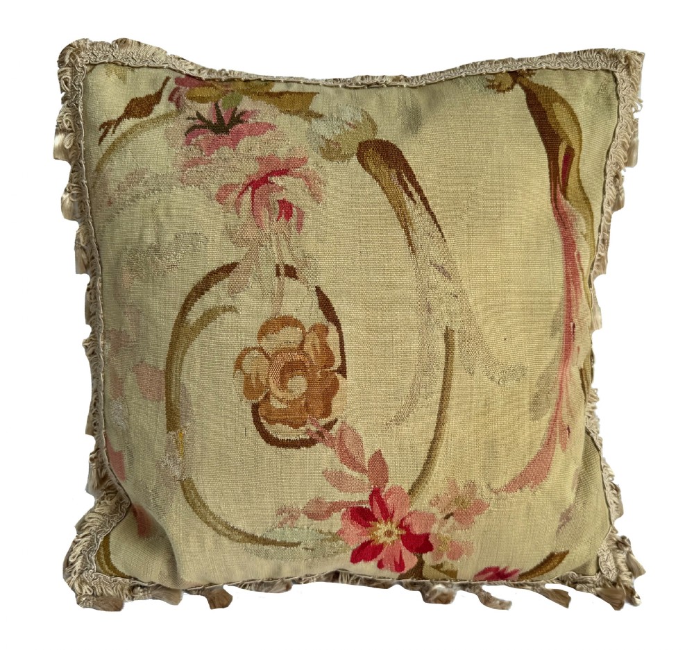 A square antique French Aubusson tapestry cushion featuring soft pink floral motifs and scrolling foliage with a fringed border.