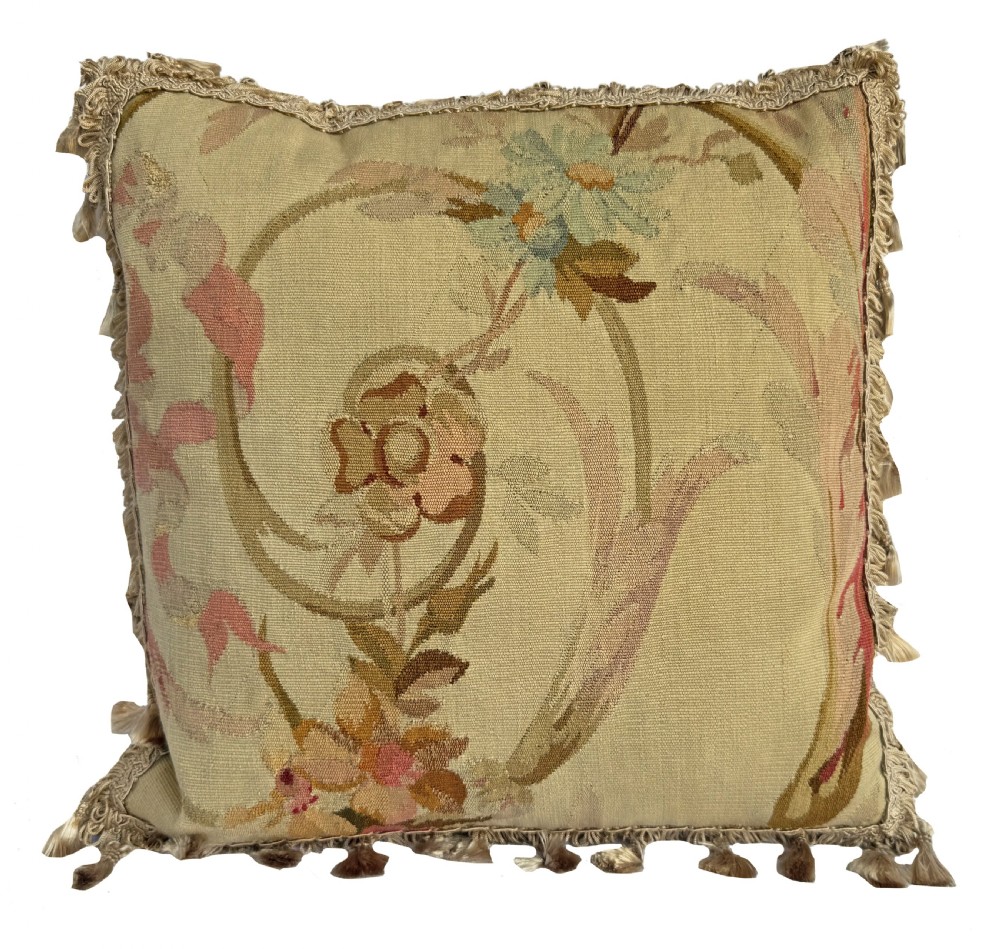 A square flat-weave tapestry cushion featuring a central floral scroll motif in pastel tones with fringe edging on a cream background.