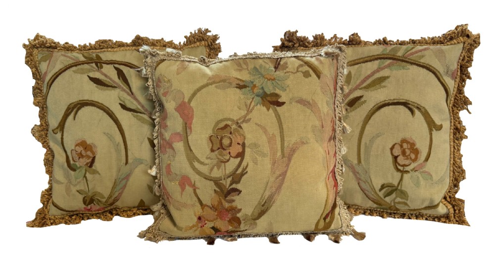 A square flat-weave tapestry cushion featuring a central floral scroll motif in pastel tones with fringe edging on a cream background. View II