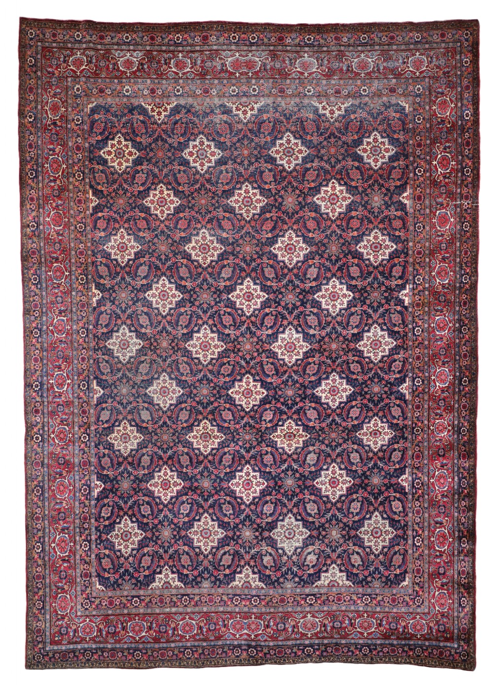 An antique Persian Kirman wool carpet featuring a repeating cream star medallion pattern on a deep navy blue central field, enclosed by intricate crimson floral borders.