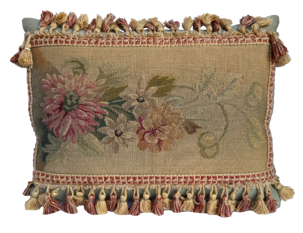 A rectangular tapestry cushion displaying a woven floral motif in beige, pink, and green tones with a fringed tassel border.