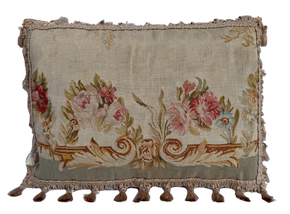 A rectangular beige tapestry cushion featuring pink floral scrollwork and tasselled edge trim.