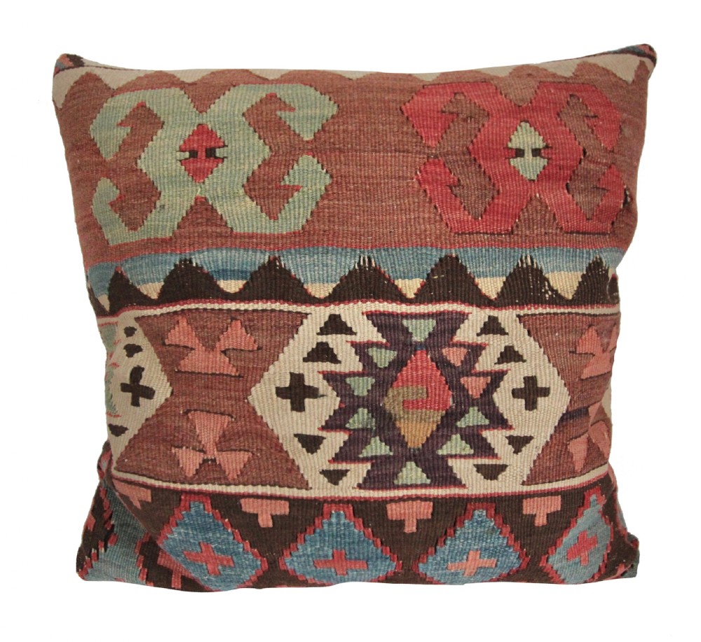 A square flat-weave wool kilim cushion featuring geometric diamond and cross motifs in terracotta red, blue, sage green, and dark brown.