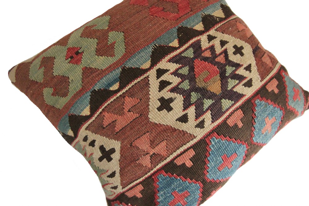 A square flat-weave wool kilim cushion featuring geometric diamond and cross motifs in terracotta red, blue, sage green, and dark brown. View III
