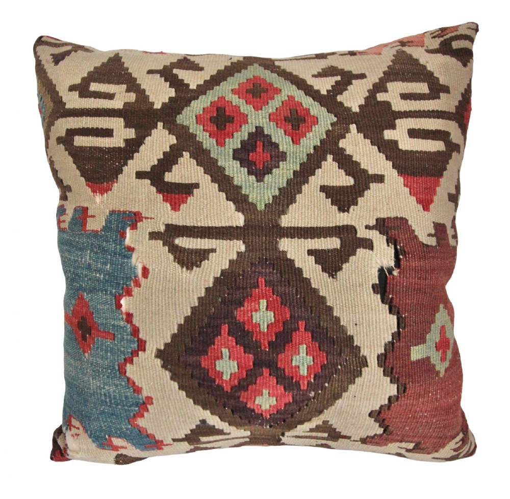 A square scatter cushion crafted from a handwoven kilim textile featuring geometric diamond motifs in dark brown, red, teal, and soft green on a cream background.